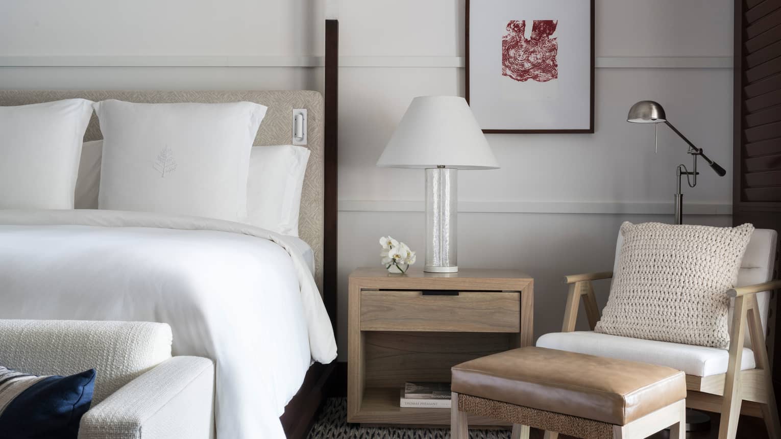 Close-up of white hotel bed, nightstand with white lamp, modern armchair and leather footstool