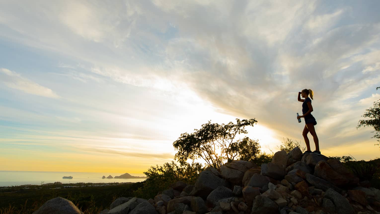 Silhouette of a woman standing on a rock with a water bottle in hand as the sun sets beyond the ocean