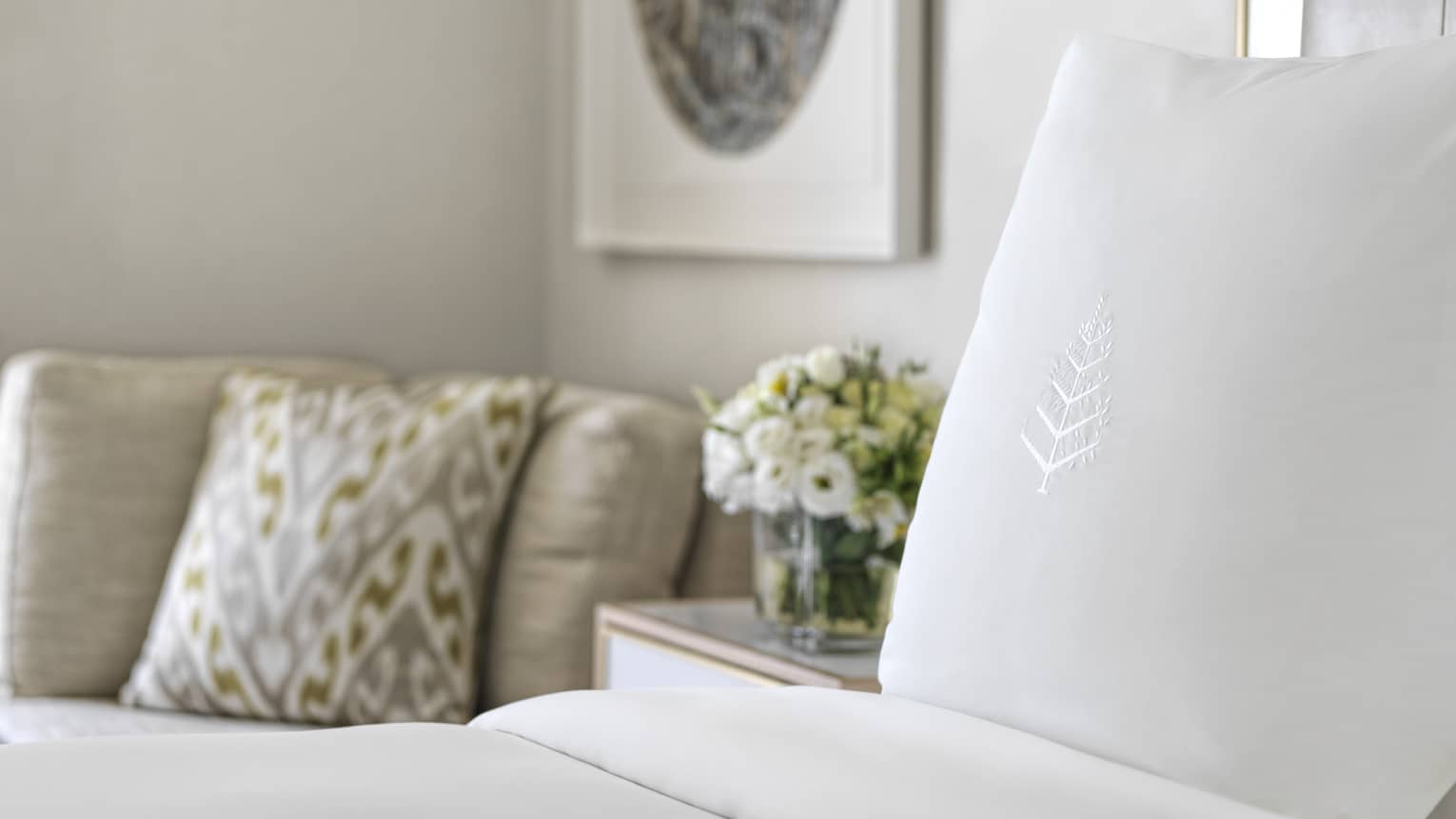 White Four Seasons–embroidered pillowcase on bed, flowers on nightstand, beige chair with pillow in corner