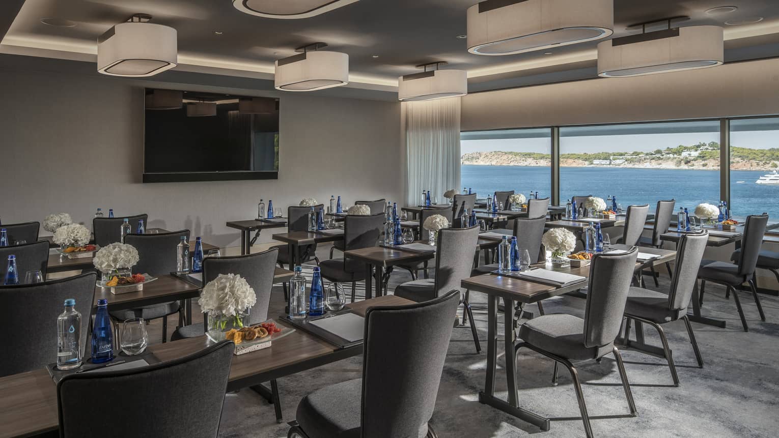 Rows of desks with water, flowers, snacks and grey chairs facing TV with ocean view