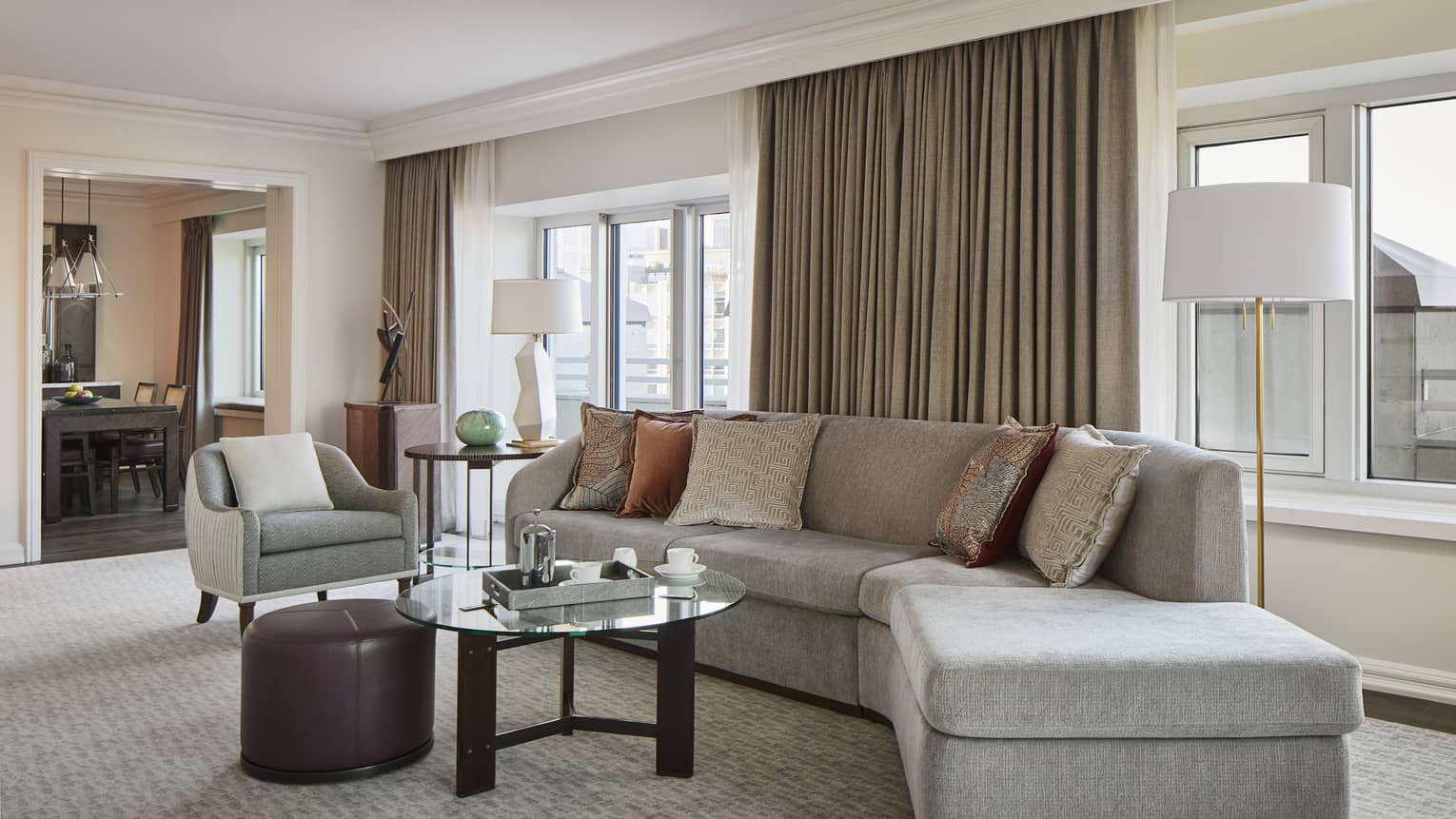 Grey sectional sofa, throw pillows, round coffee table, brown leather ottoman, club chair, view into dining area
