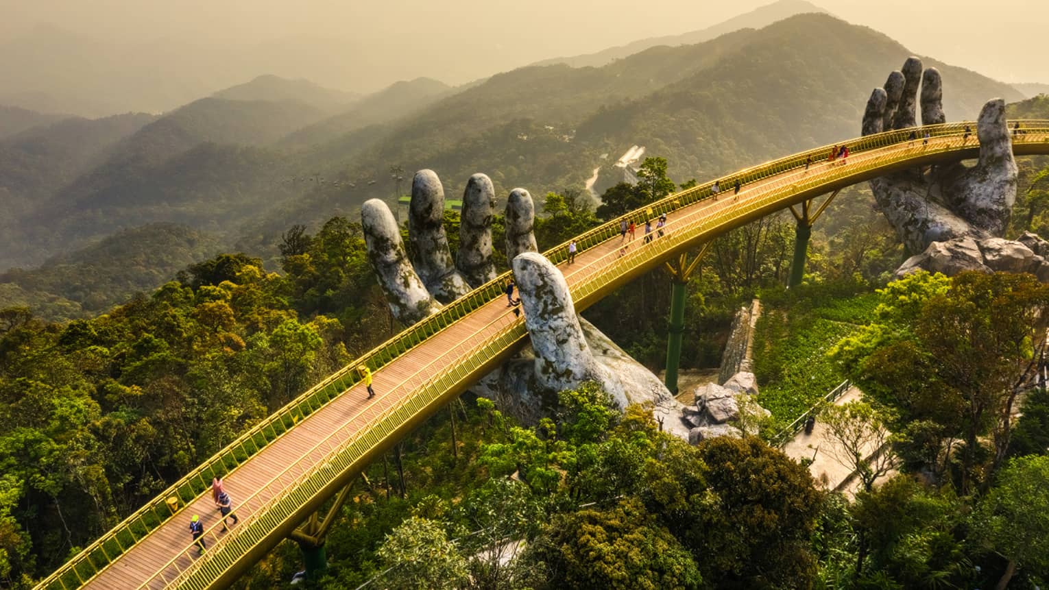 Aerial view of people on a wood-slat bridge above a forest canopy, with golden railings supported by two giant stone hands.