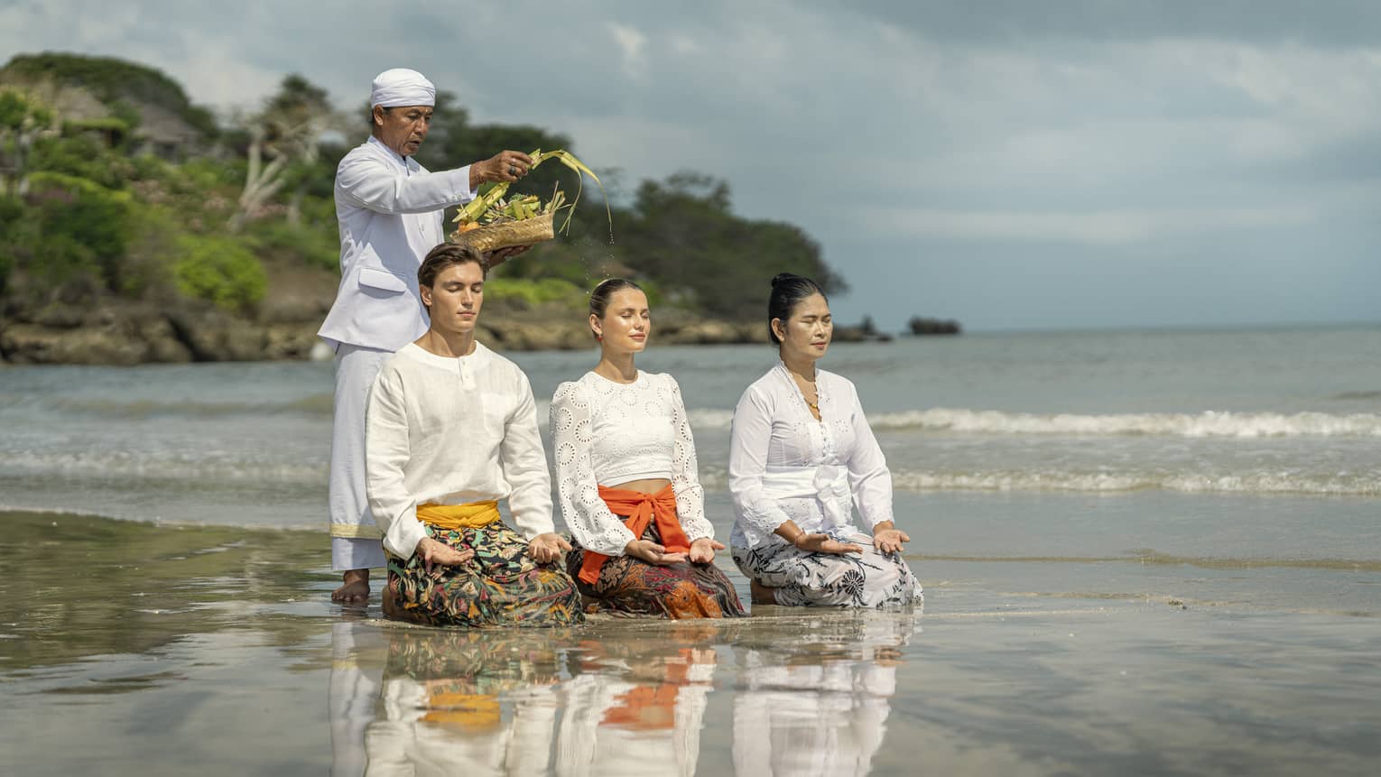 Three adults sit in shallow water with palms up on their knees as another sprinkles water from palm fronds on their heads.
