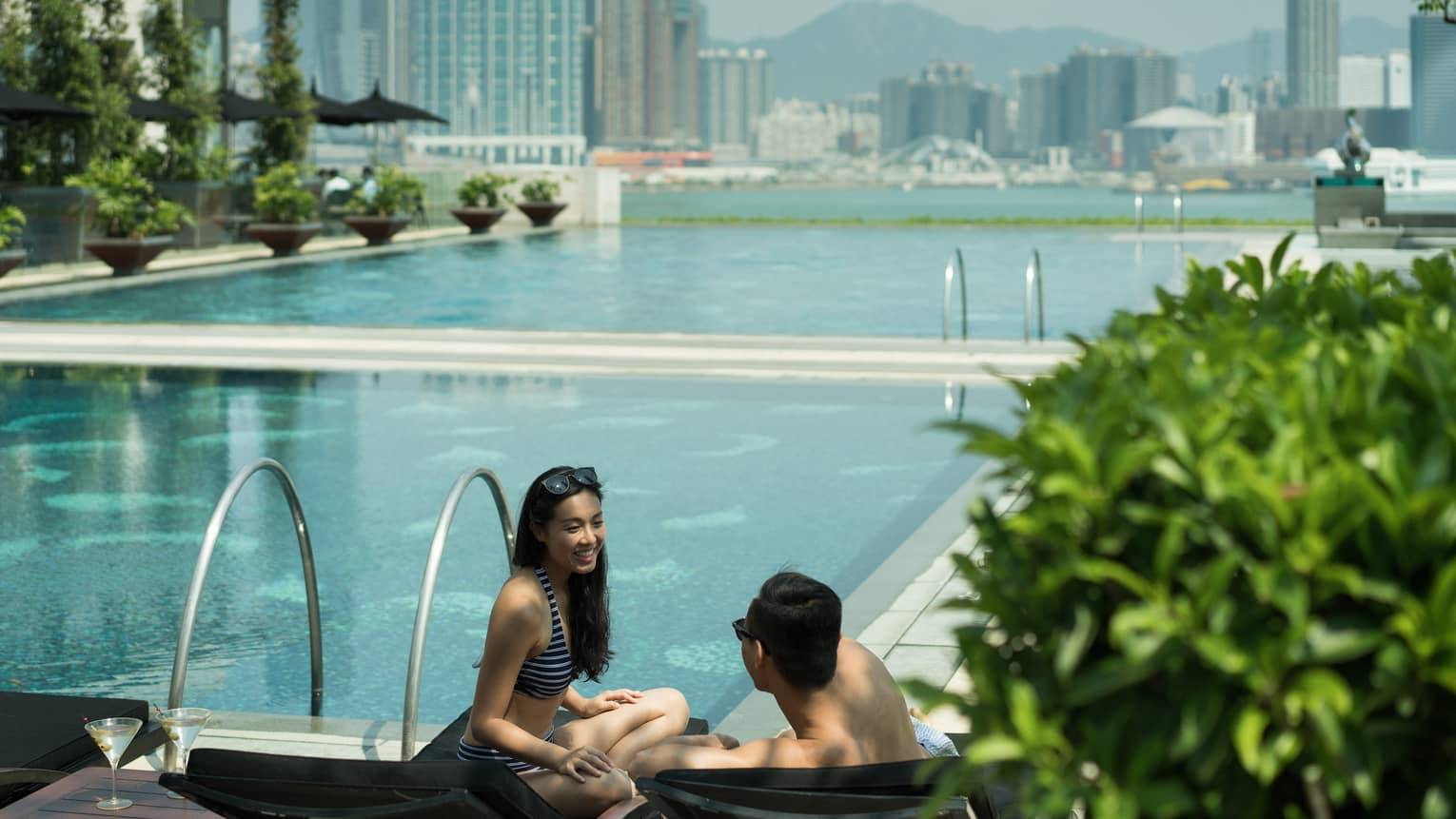Two people are by the pool edge, and the city skyline is behind them. The pool area has greenery, lounge chairs and plants and there is a clear sky overhead.