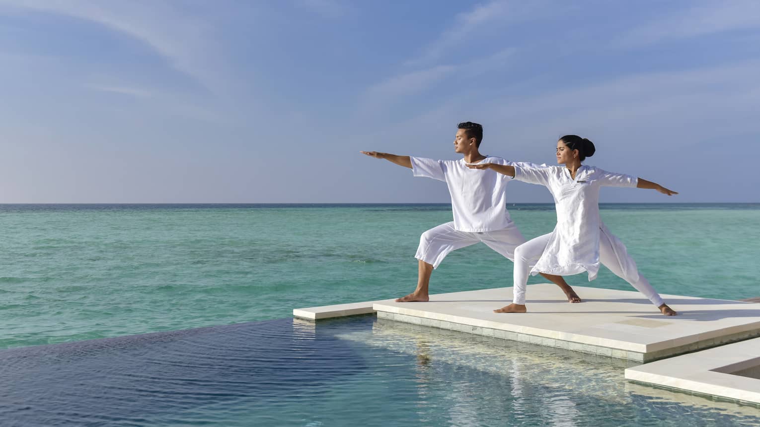 A couple dressed in all white does yoga on a white dock over the sea