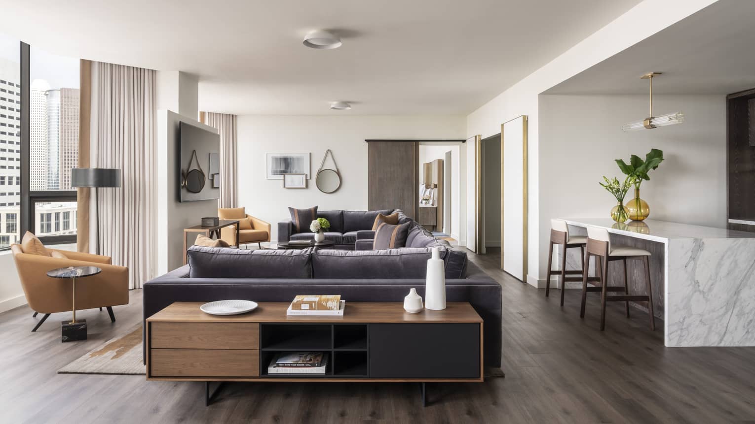 The penthouse suite, featuring a dark sofa, a brown leather chair, an island with two bar stools, a shelf unit, and large windows to the side.
