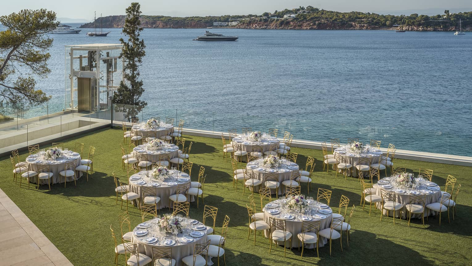 Ten round tables with gold and white chairs and flowers outside on grass overlooking ocean