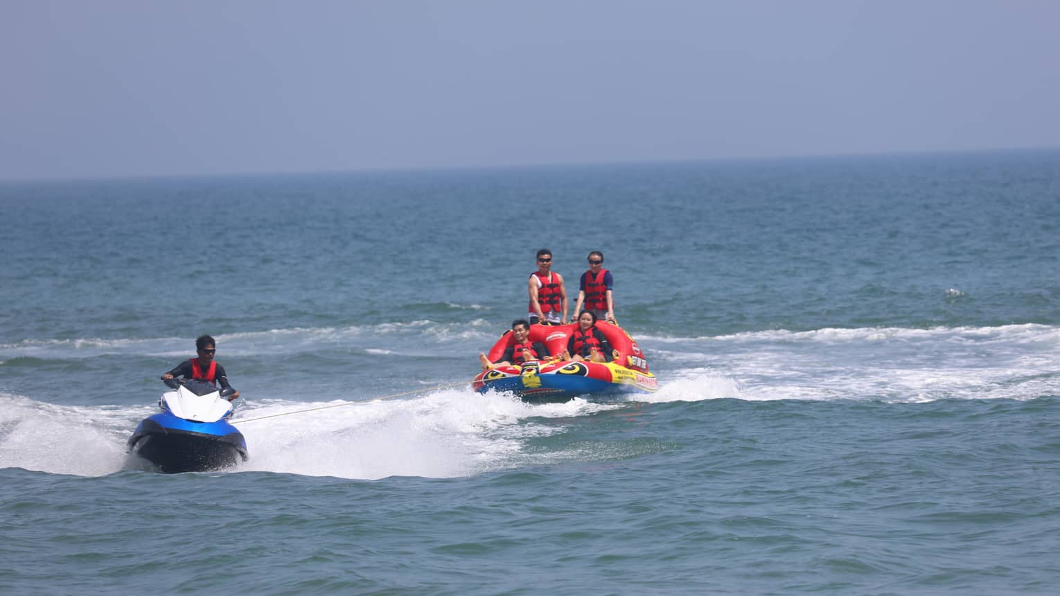 Four people with life jackets in dingy, pulled by man riding jet-ski 