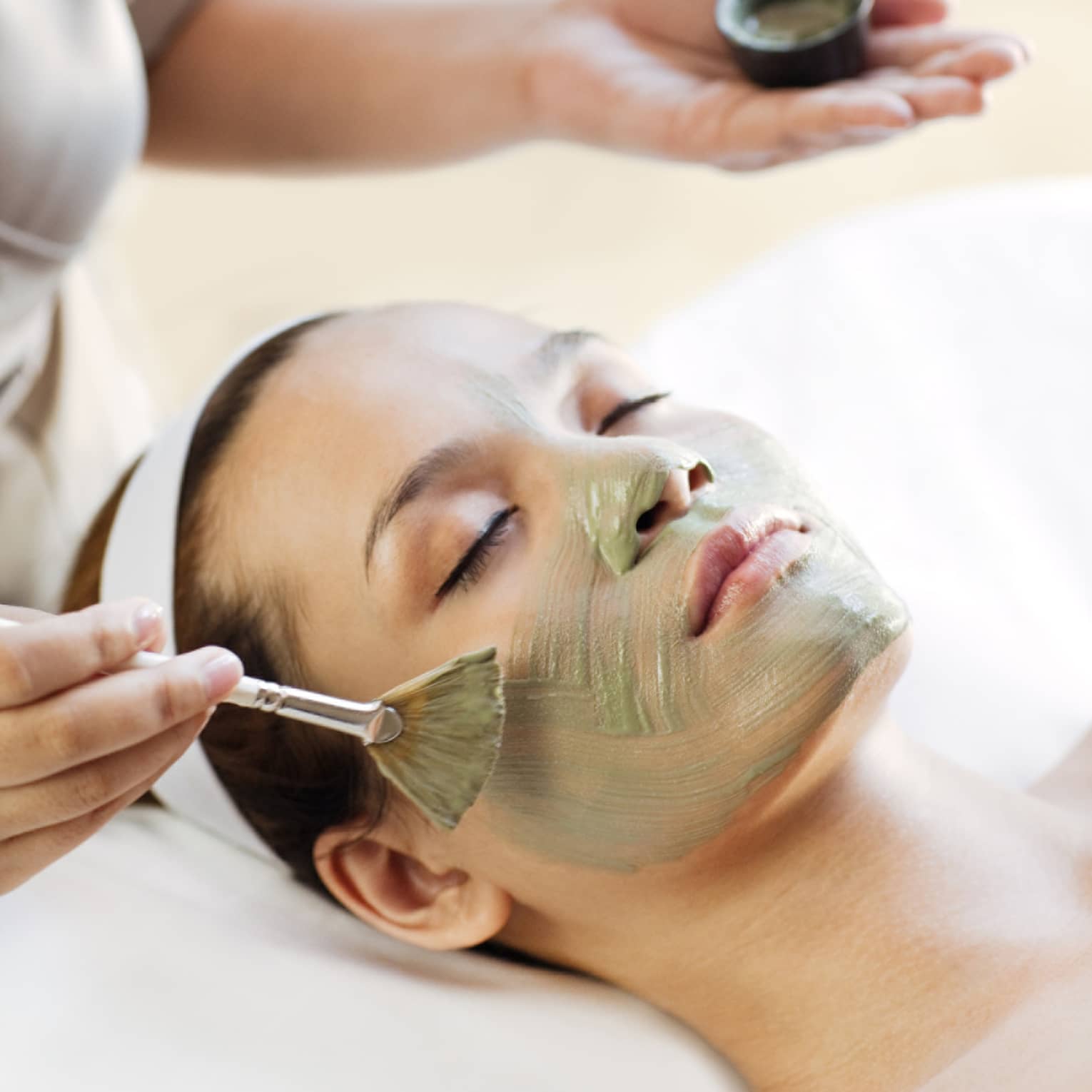 Spa staff holding brush paints green mask on woman's cheeks, chin, nose 