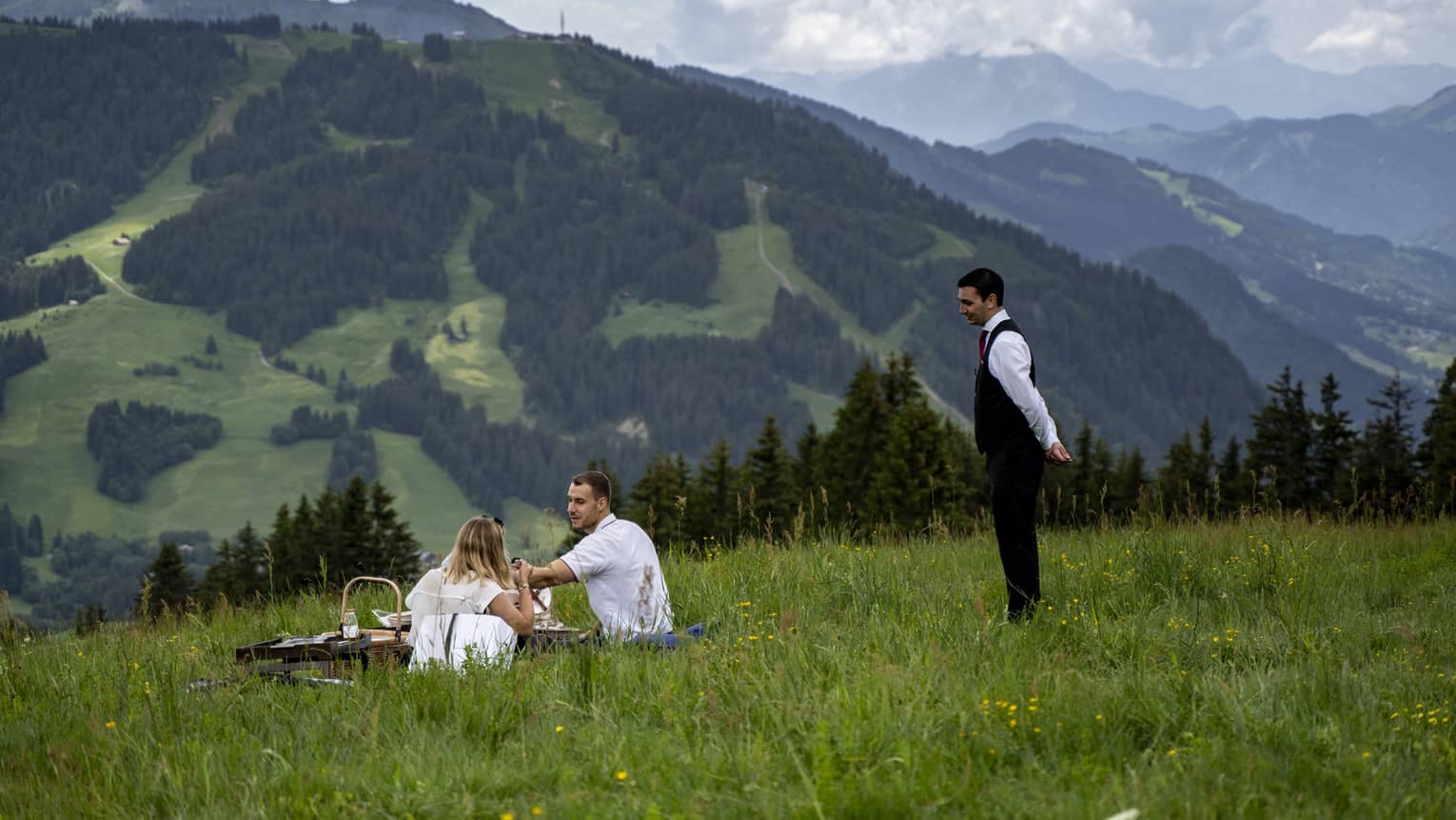 Man and woman sit on blanket with picnic on lush green mountainside, server assisting