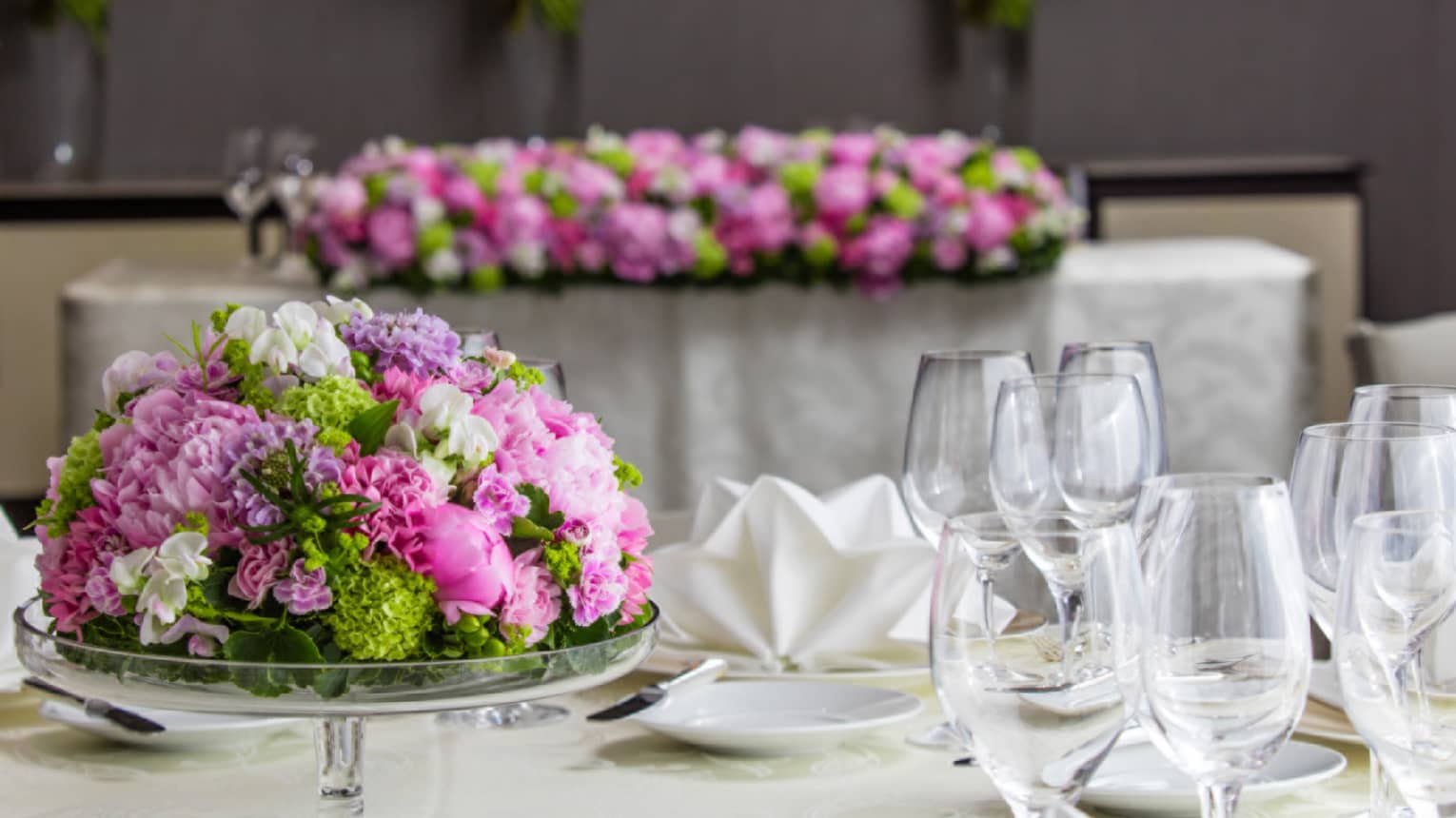 Pink and green floral arrangement in glass cake stand on table with glassware