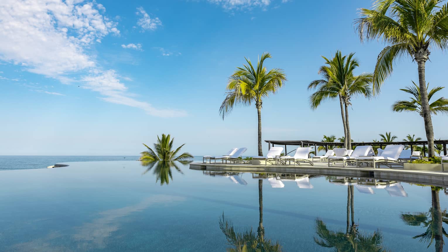 A calm infinity pool reflects the blue sky, clouds and palm trees