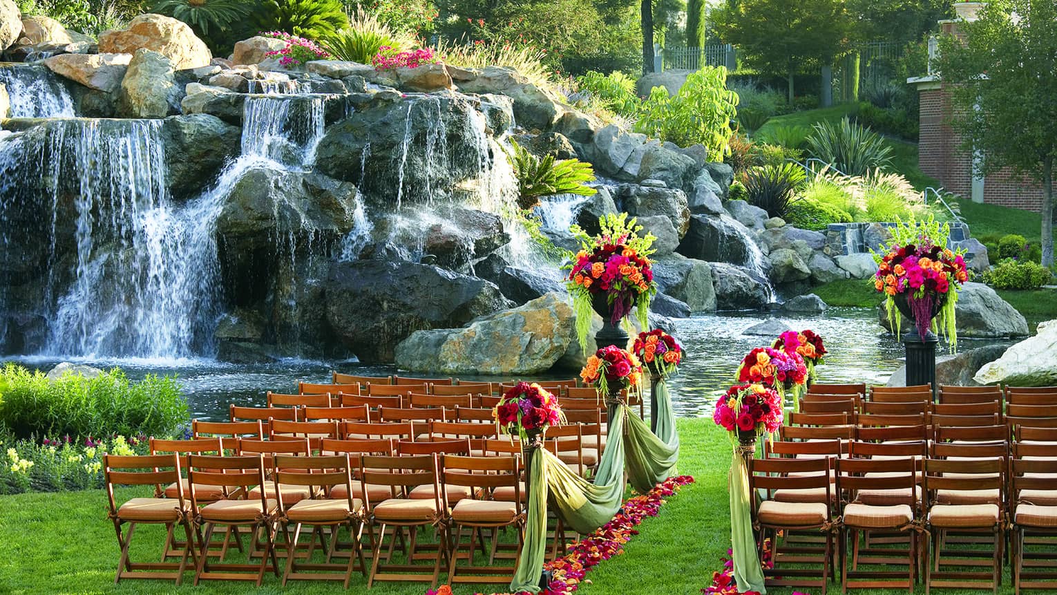 Rows of chairs face waterfall lawn, draped with green material, brightly coloured flowers, petals on lawn 