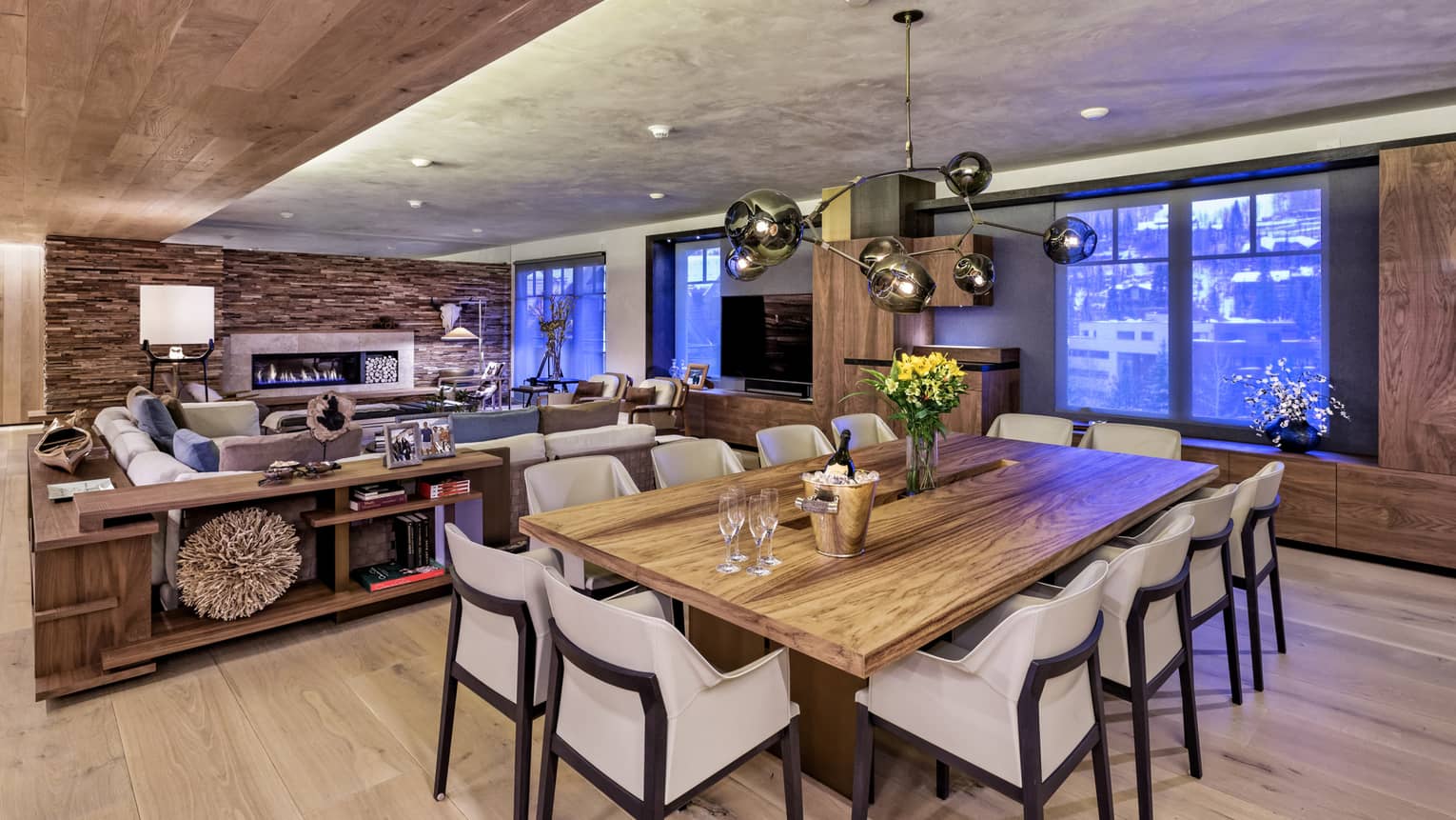 Modern wood farm-style dining table lined with white chairs, Champagne on ice, in front of living room