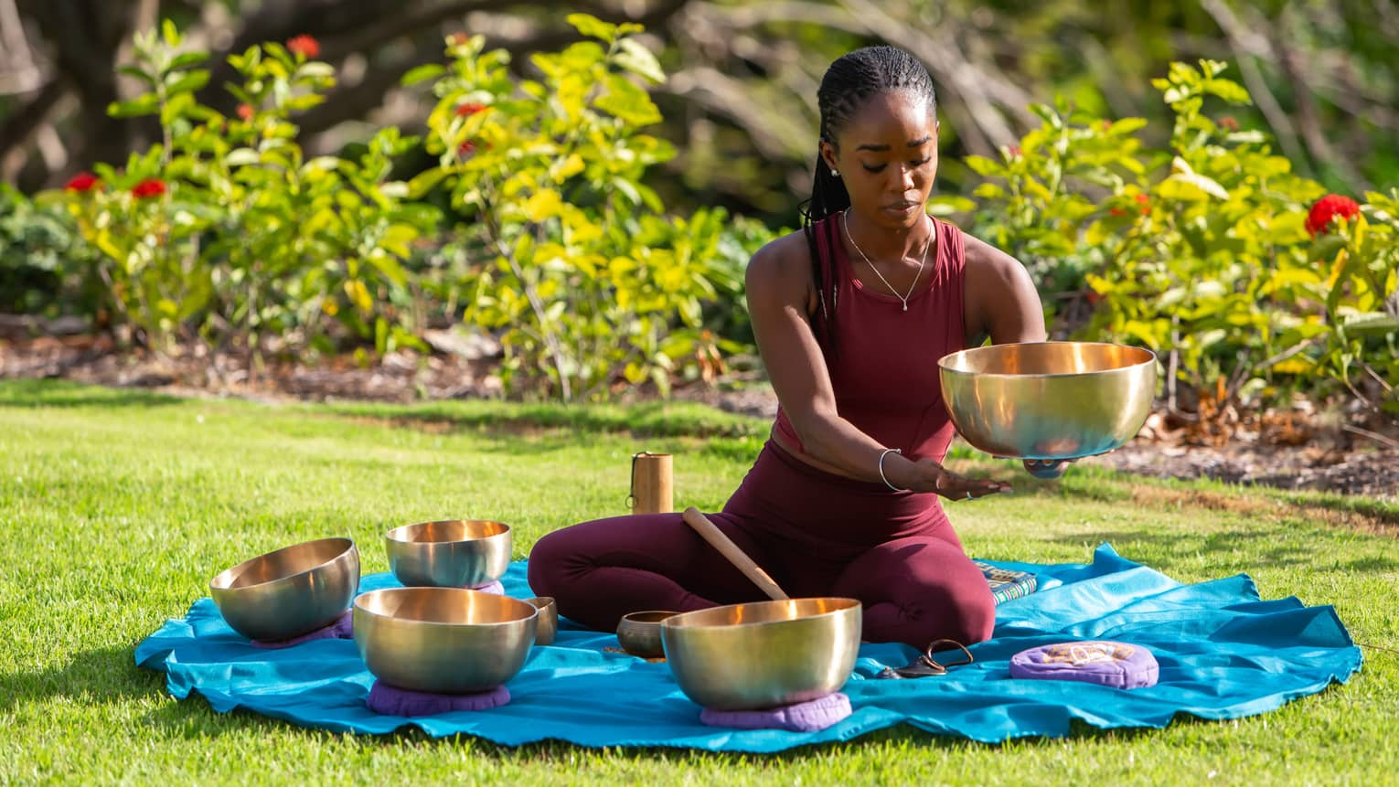 A sound bath practitioner holds a brass singing bowl and gazes downward while sitting on a mat amid various instruments.