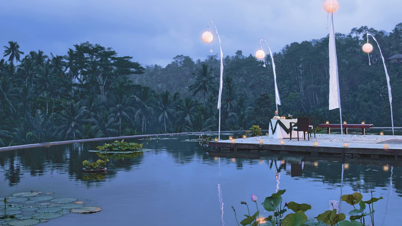 Round white paper lanterns hang from poles over candle-lit dining table on wood platform over lotus pond at dusk