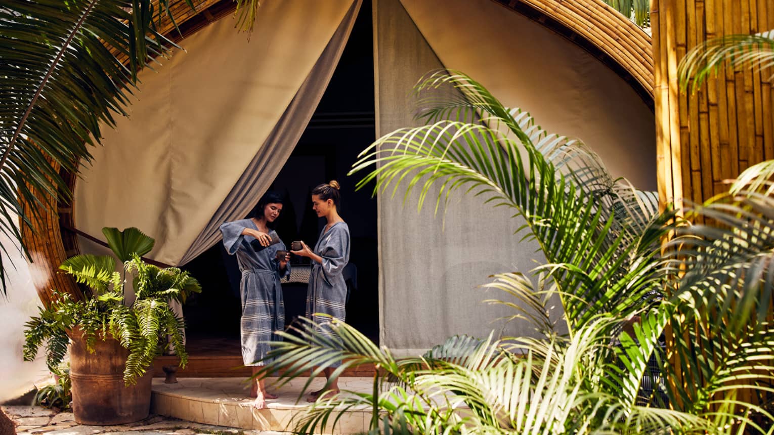 Two women in spa robes entering a spa tent surrounded by palm trees.