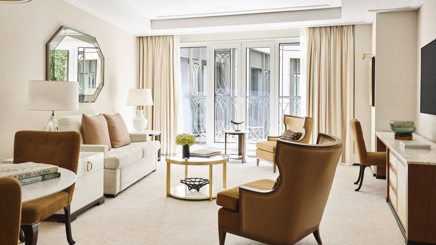 Luxury hotel suite with cream sofa, gold-accented coffee table, tan armchairs, octagonal mirror, TV and floor-to-ceiling windows with ornate iron railings