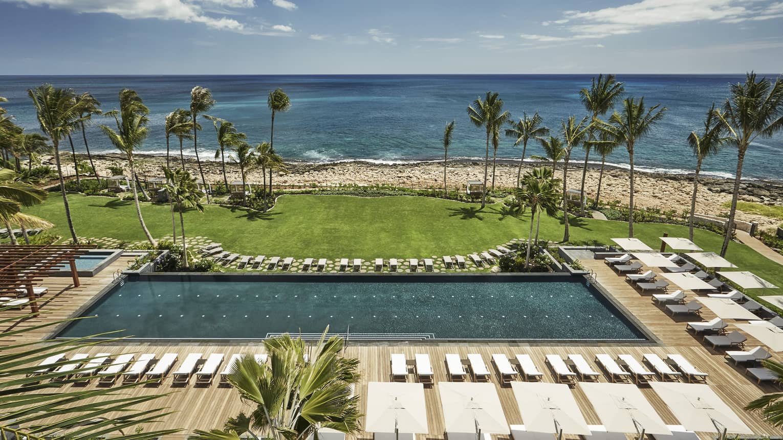 Aerial view of large outdoor swimming pool lined with white deck chairs, lawn, beach