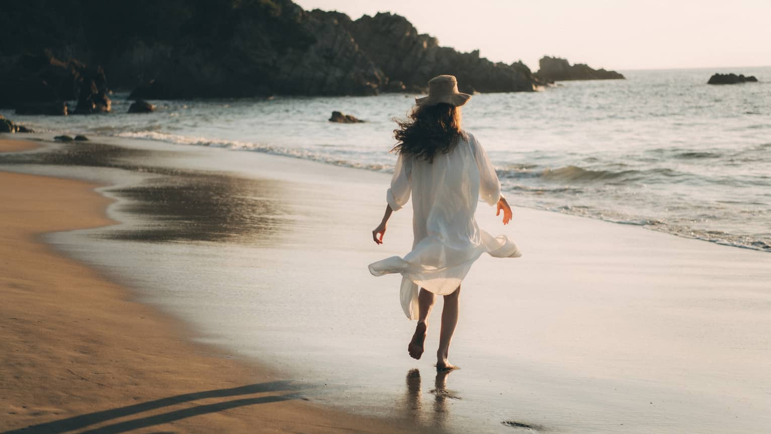 A woman in a long white shirt walking along a beach.