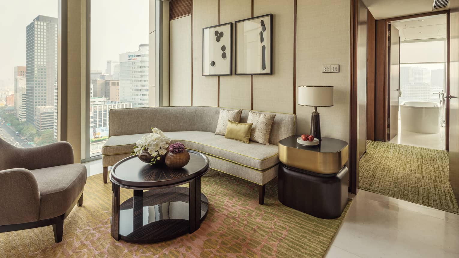 City-View Executive Suite curved sofa under window, armchair, table with lamp by hallway to bathroom