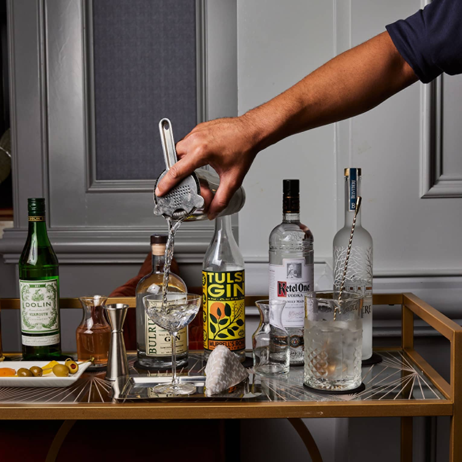 A bartender pours from a shaker into a coupe glass next to bottles of gin, vodka and vermouth, a jigger, carafes and olives.