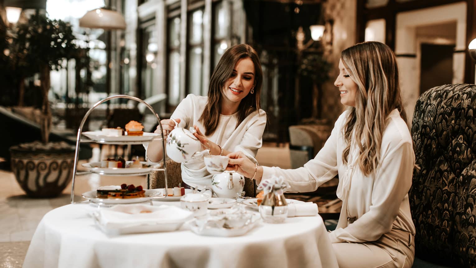 Two women in neutral outfits seated, smiling over afternoon tea in hotel lobby bar