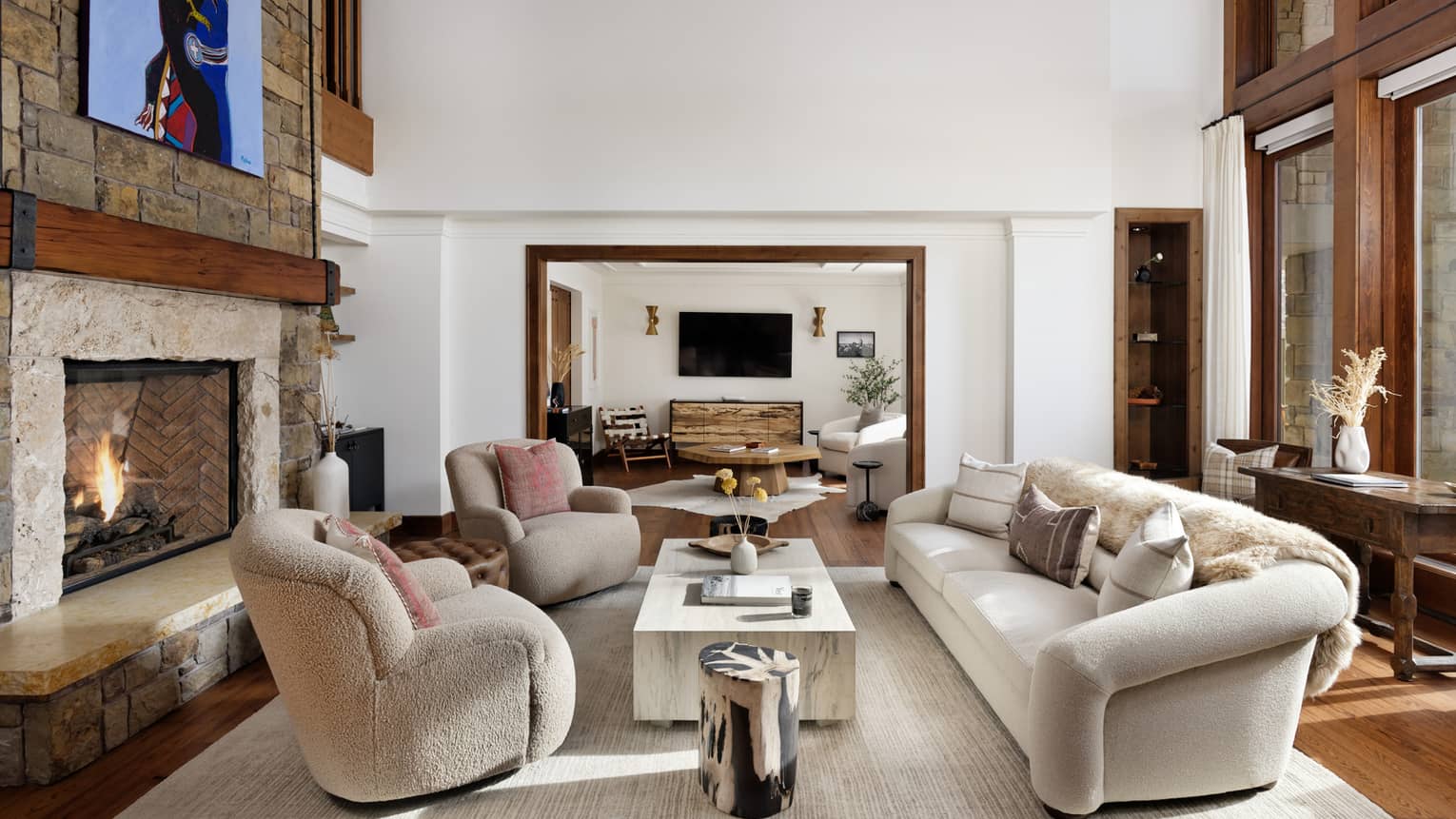 Mountain residence living room with a stone fireplace, sofa and armchairs around a coffee table, leading into a connecting seating area with a TV on the wall