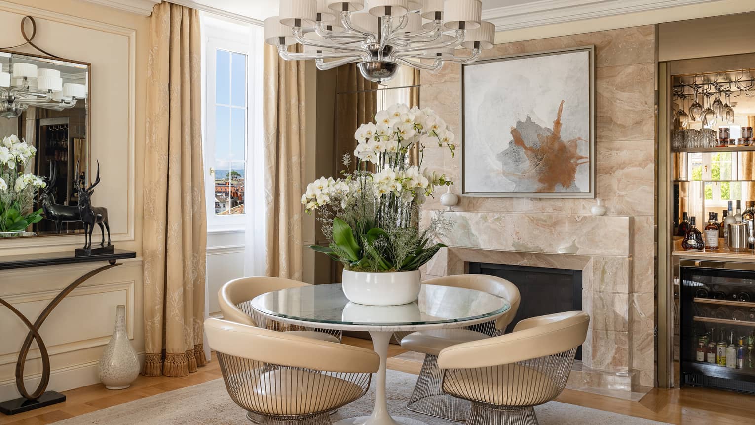 Luxury suite dining area with a round table, beige chairs, floral arrangement, chandelier, fireplace, bar, mirror and decorative sculpture on a console table
