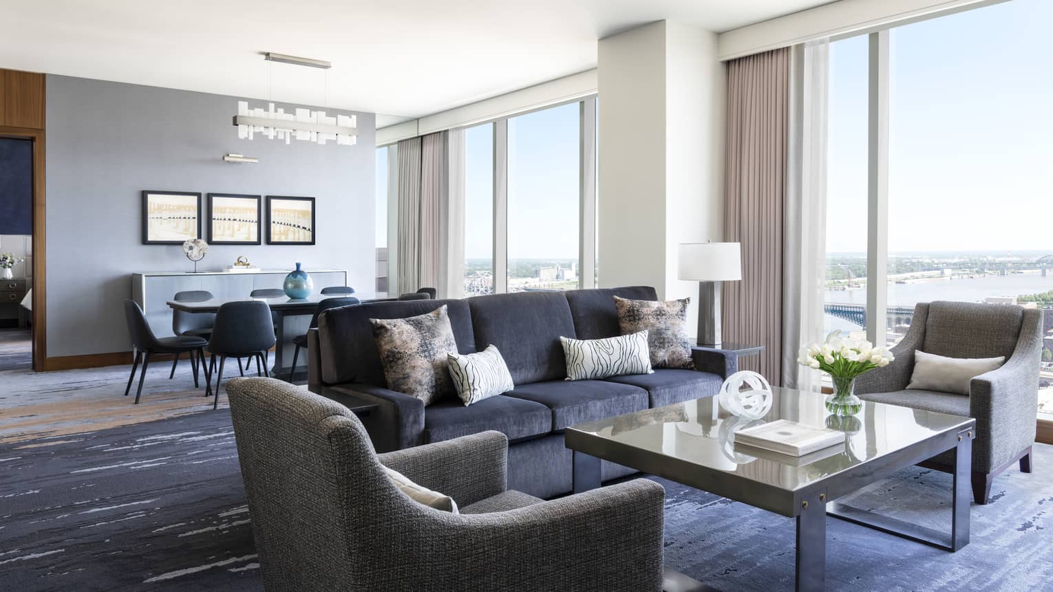 Hotel suite living and dining area with large windows offering a river view, dark gray sofa, armchairs, a coffee table and a dining table with modern chairs