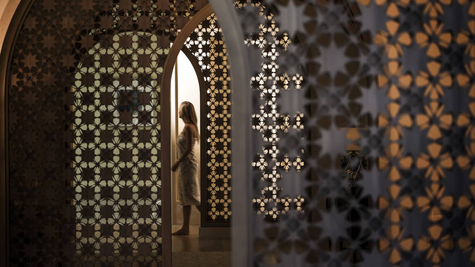Woman in white wrap walks through dimly-lit decorative entranceway in hall