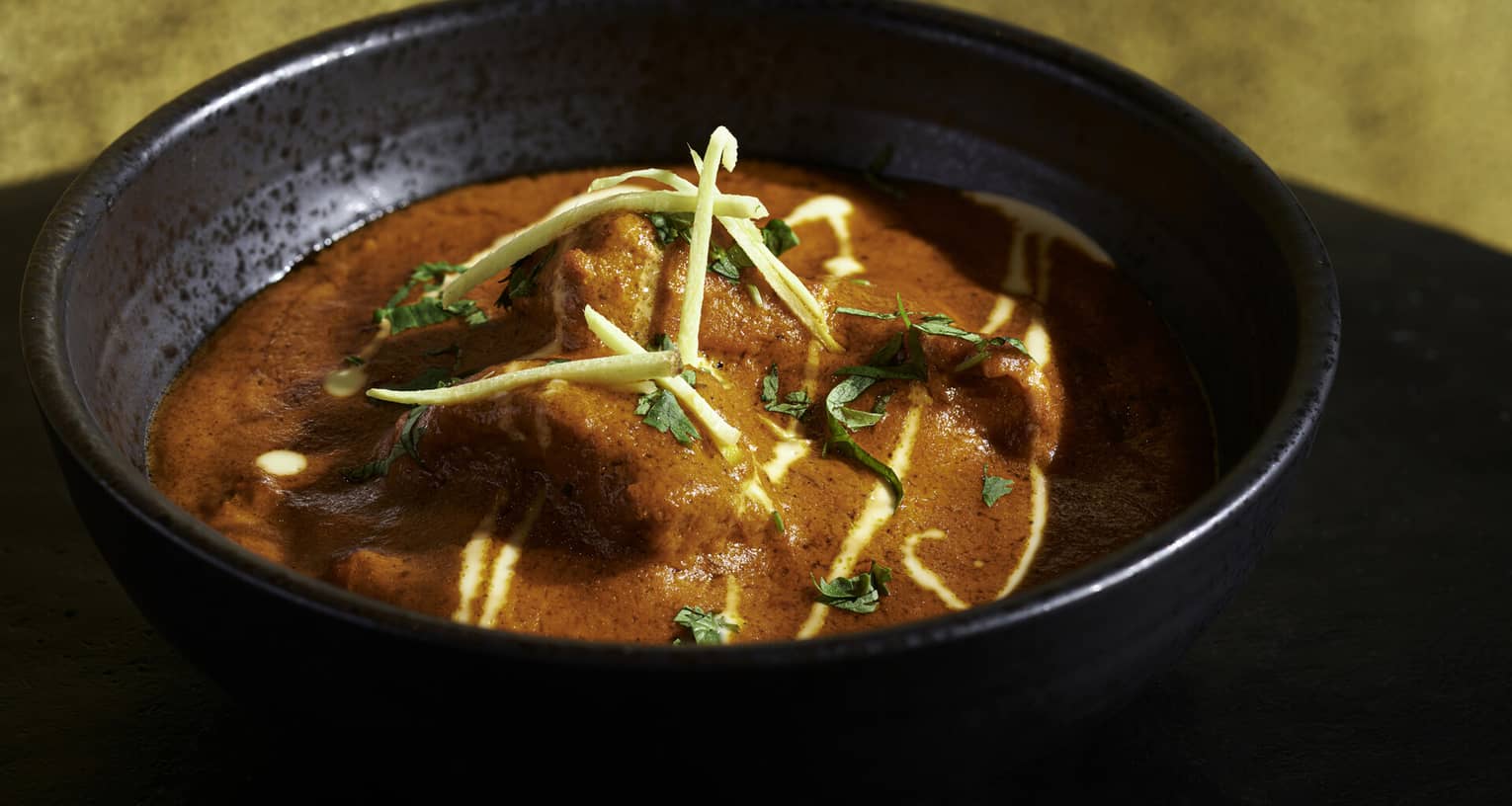Bright red Tandoori dish with chicken, fresh herb garnish in black earthenware bowl