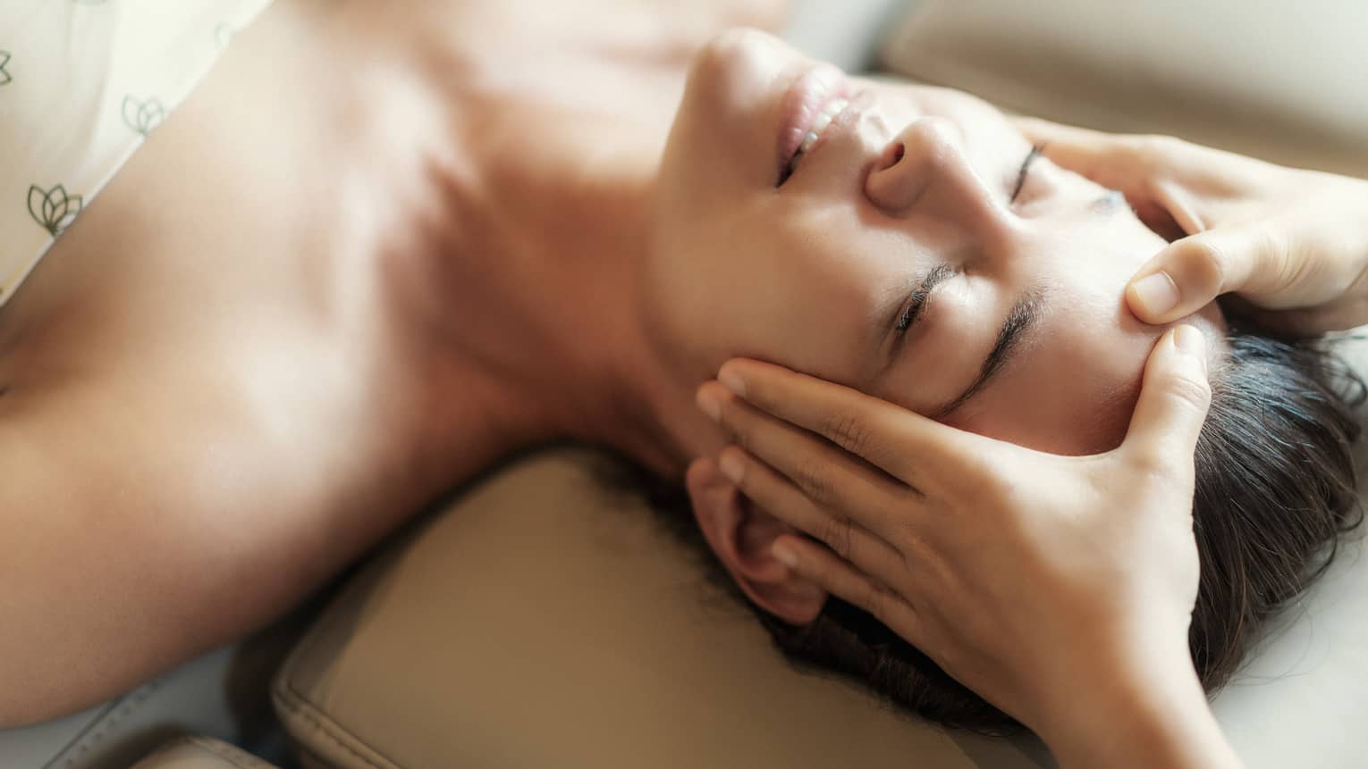 Spa facial, two hands rest on woman's forehead as she closes her eyes, lays under sheet on treatment table