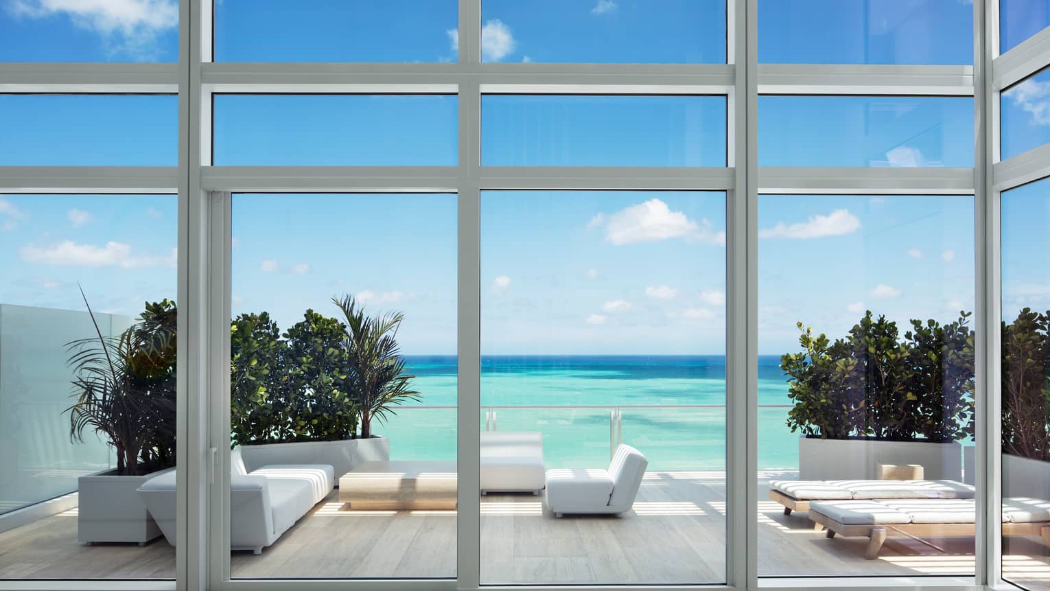 View from glass walls to large patio with modern white furniture, turquoise ocean