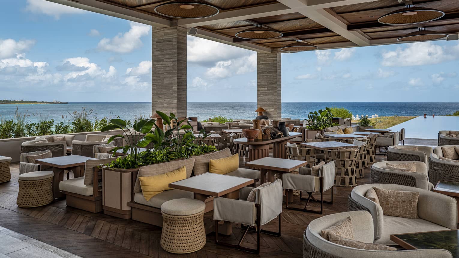 A spacious, covered outdoor dining space with small tables, comfortable chairs, plantings and ocean views