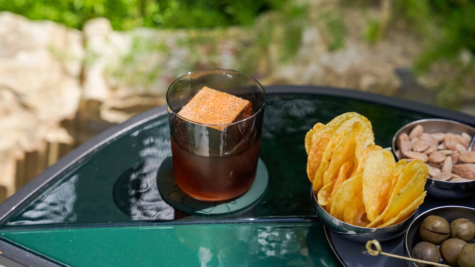 Dark-hued cocktail garnished with an orange-topped block of ice sits on a green outdoor table next to a platter of appetizers