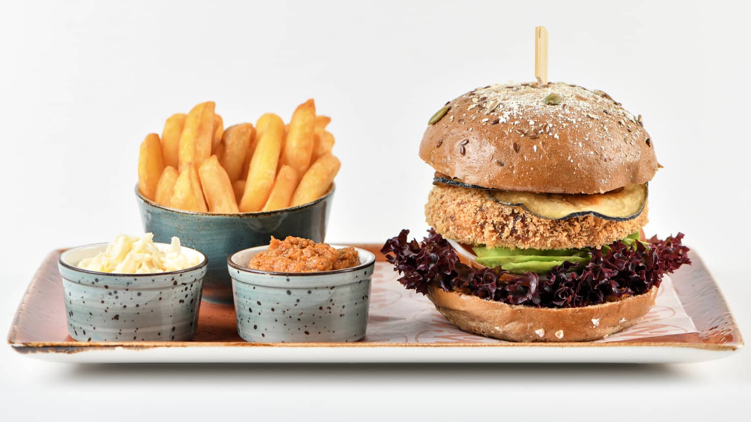 Quinoa Burger on an orange rectangular plate set next to a serving of fries and two small containers of toppings