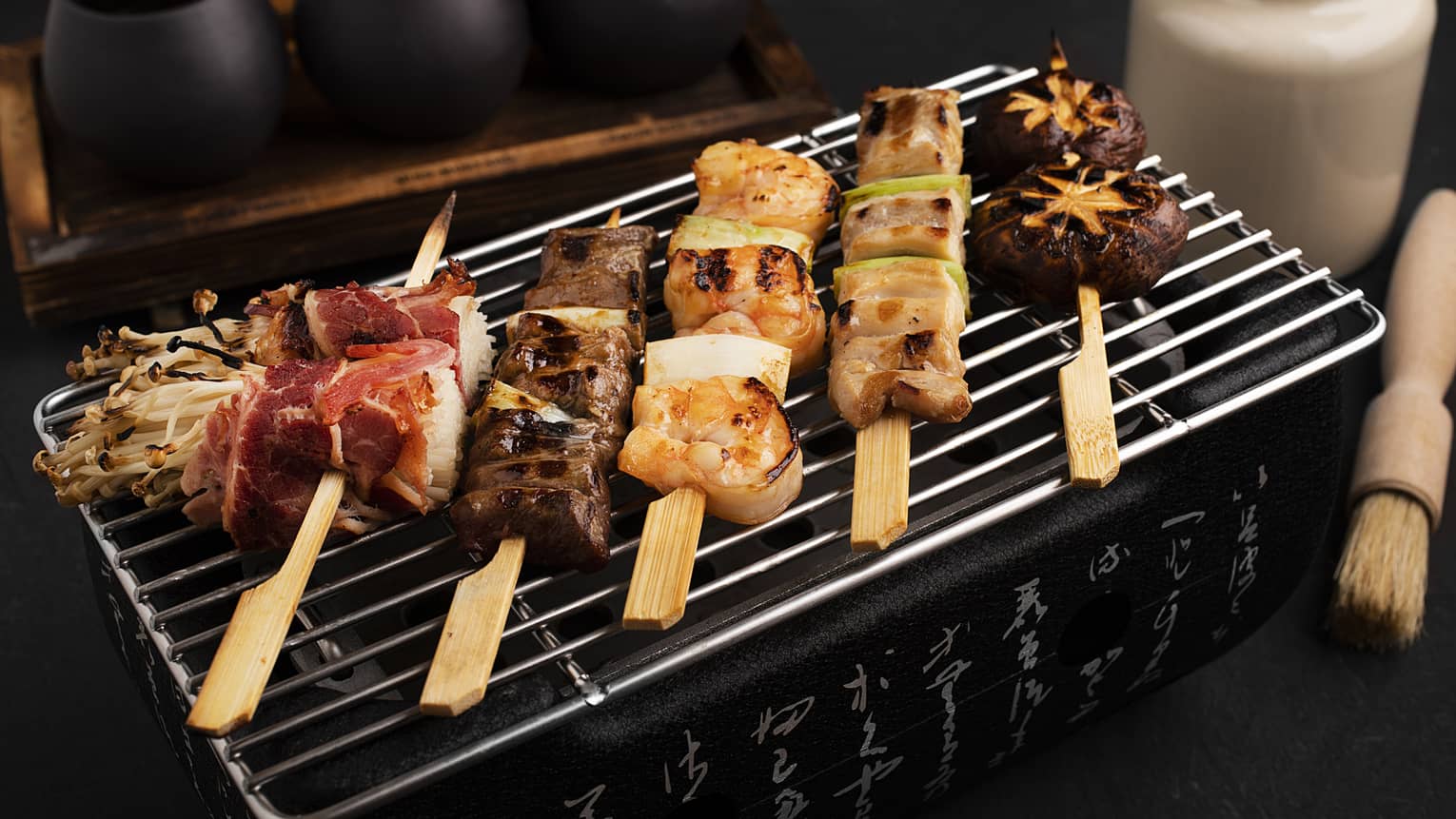 Chicken negima, beef kushiyaki, enoki bacon wraps, prawn skewers and shiitake mushrooms all on skewers laying on a grill