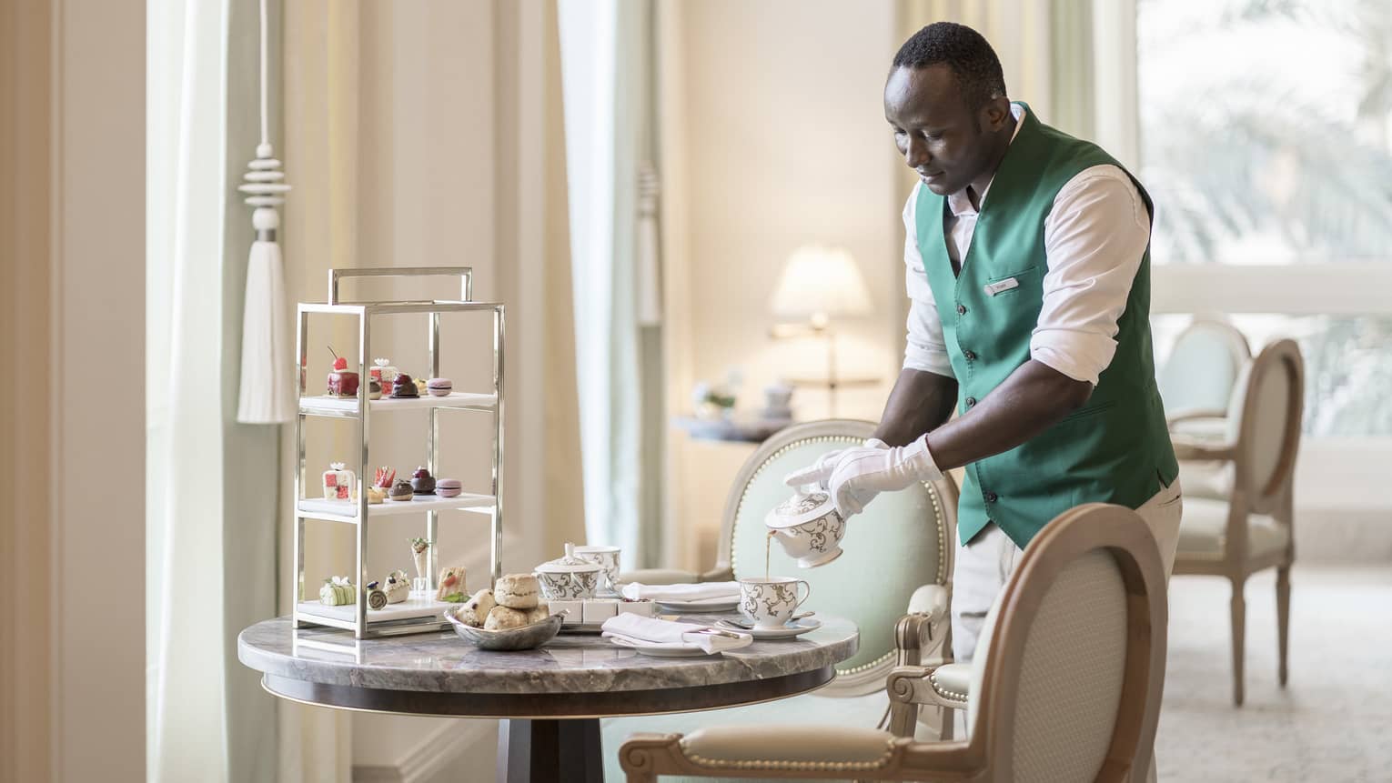 Four Seasons team member wearing a white shirt and green vest pours a cup of tea on a table set for Afternoon Tea