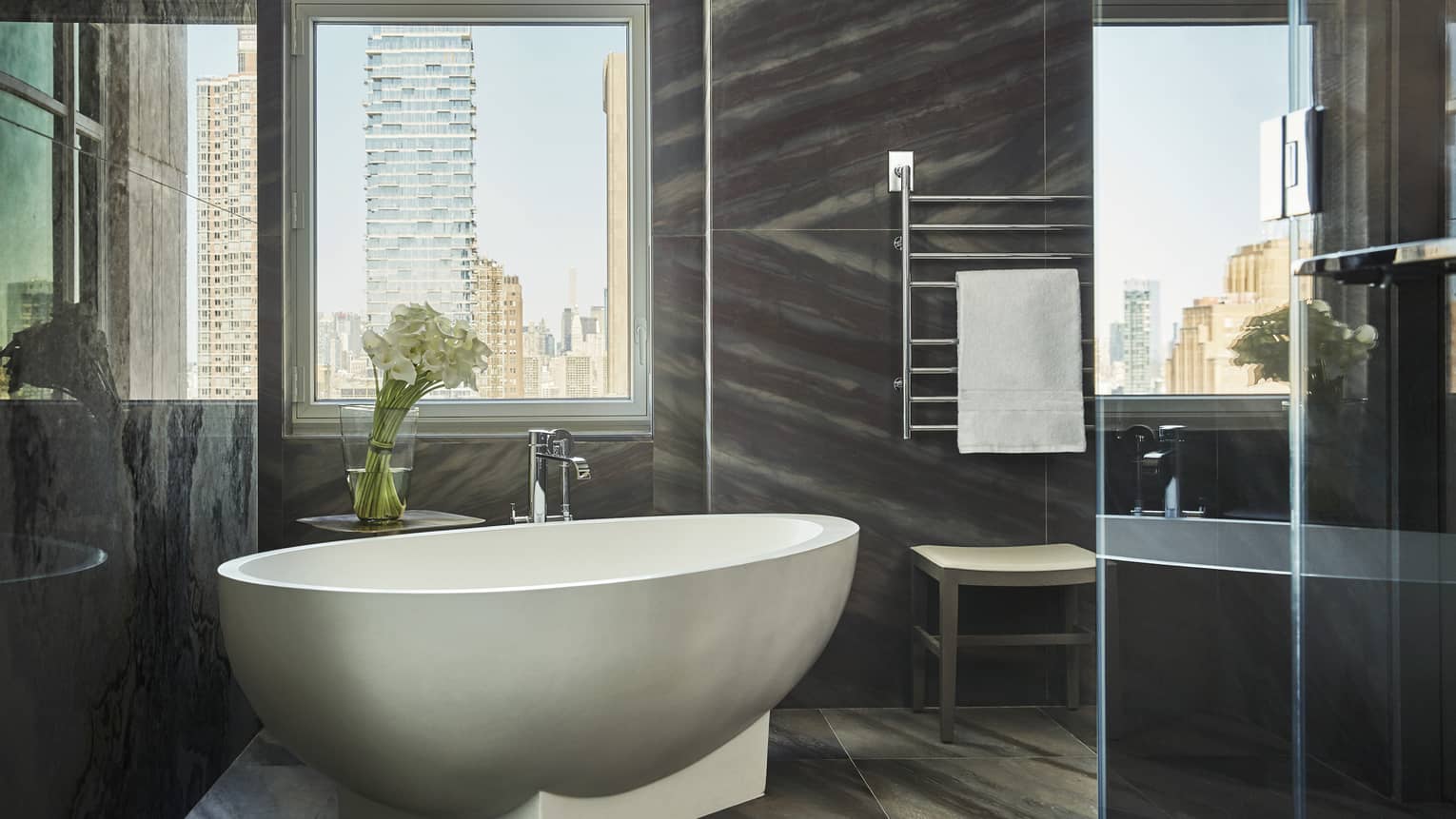 Free-standing white bathtub under tall window, modern grey tile bathroom