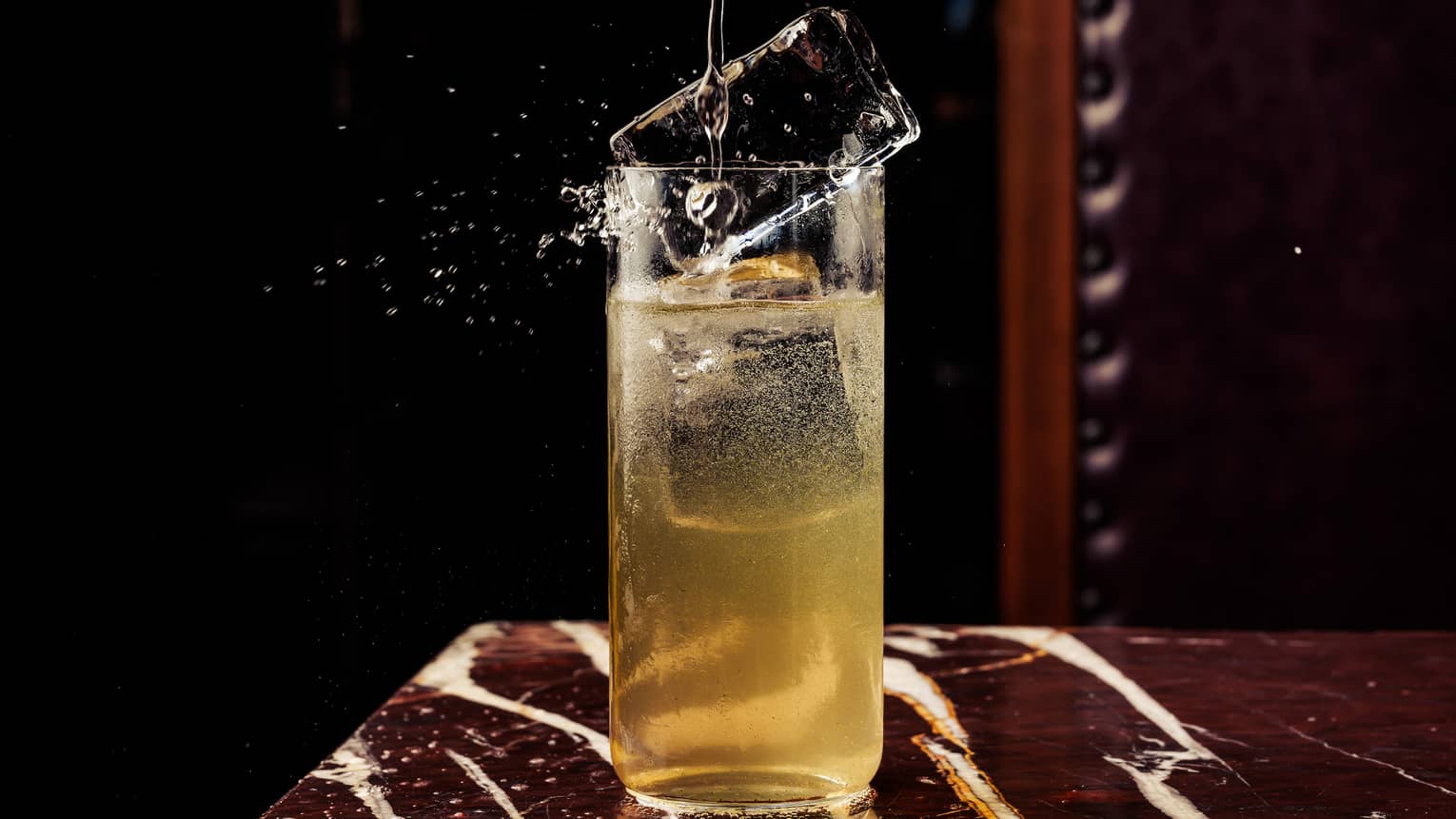 A large rectangular ice cube splashes down into a high-ball glass filled almost to the top with a gold-colored beverage