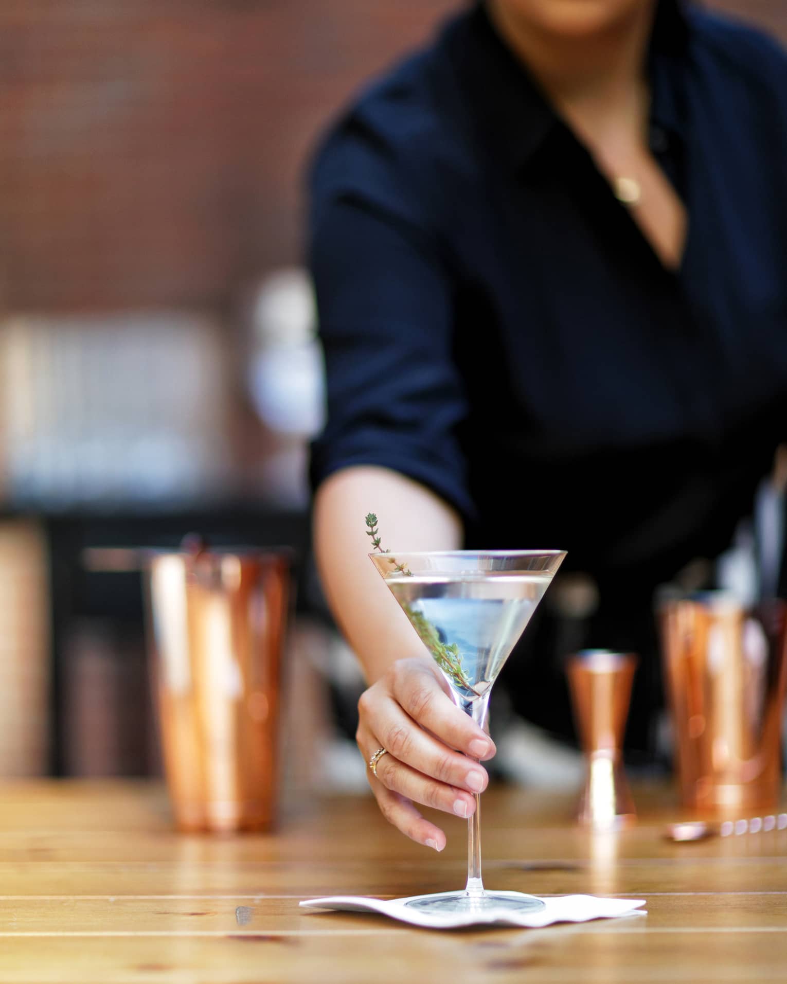 Bartender offering a martini garnished with a sprig of fresh thyme set on a small napkin, cocktail shakers in the background.
