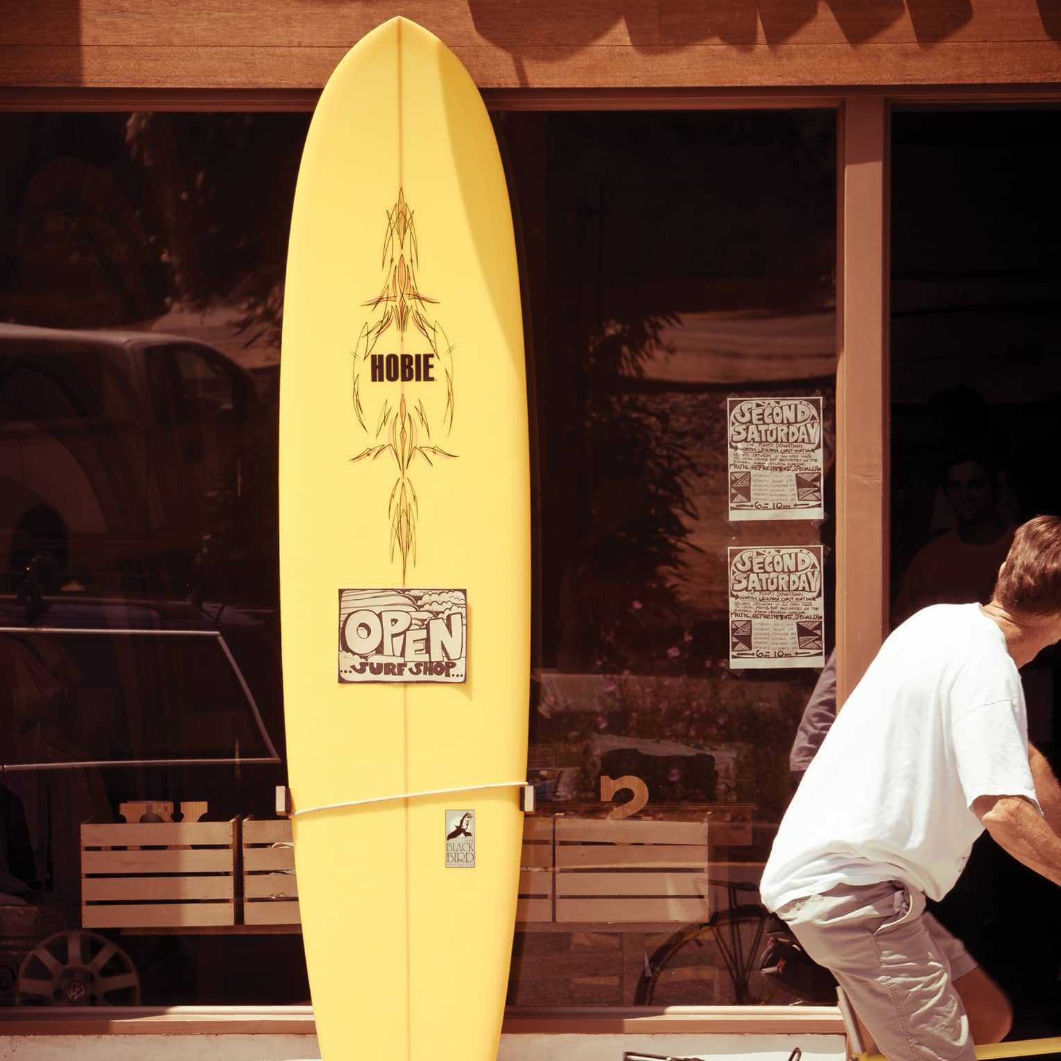 In the sun’s warm glow, a cyclist turns their head while riding by a yellow surfboard resting against a surf shop’s window. 