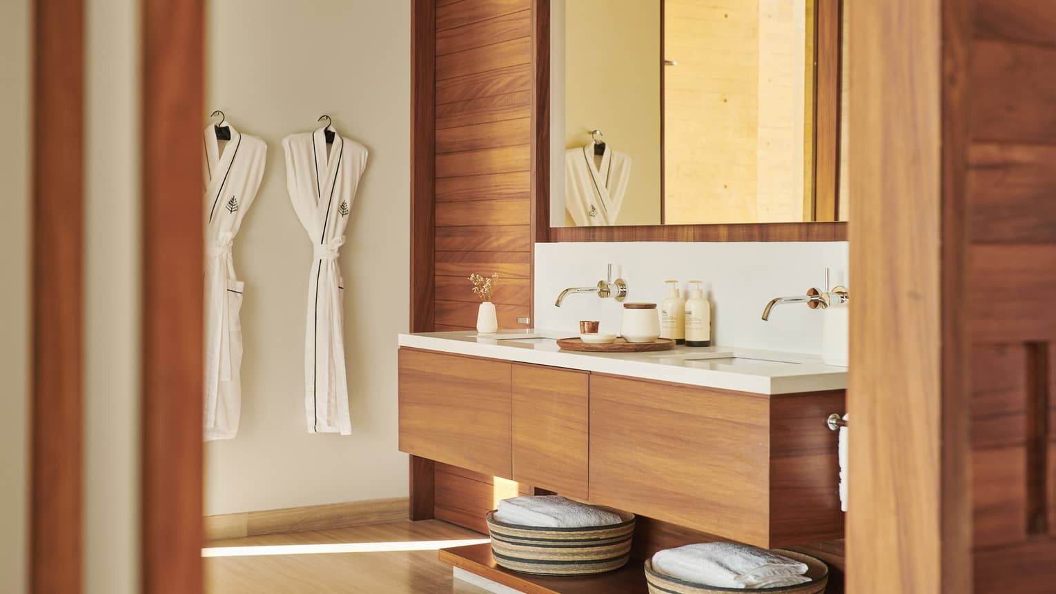 Modern bathroom with wooden accents, featuring a double-sink vanity, large mirror and neatly hung bathrobes. Baskets with towels are stored below the counter, creating a spa-like ambience.