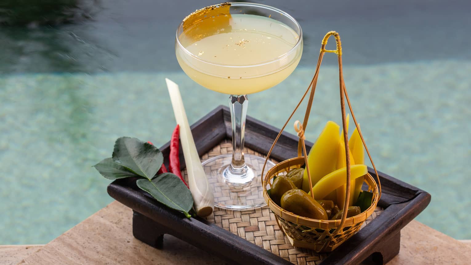 Thai cocktail on poolside tray with lime leaves, lemongrass and chilli garnishes