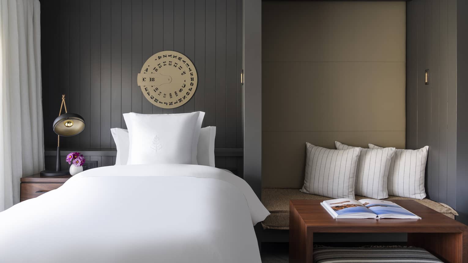 Luxury resort room with white bed, dark paneled walls, decorative wall art, bedside table with lamp and flowers and seating nook with striped pillows and book