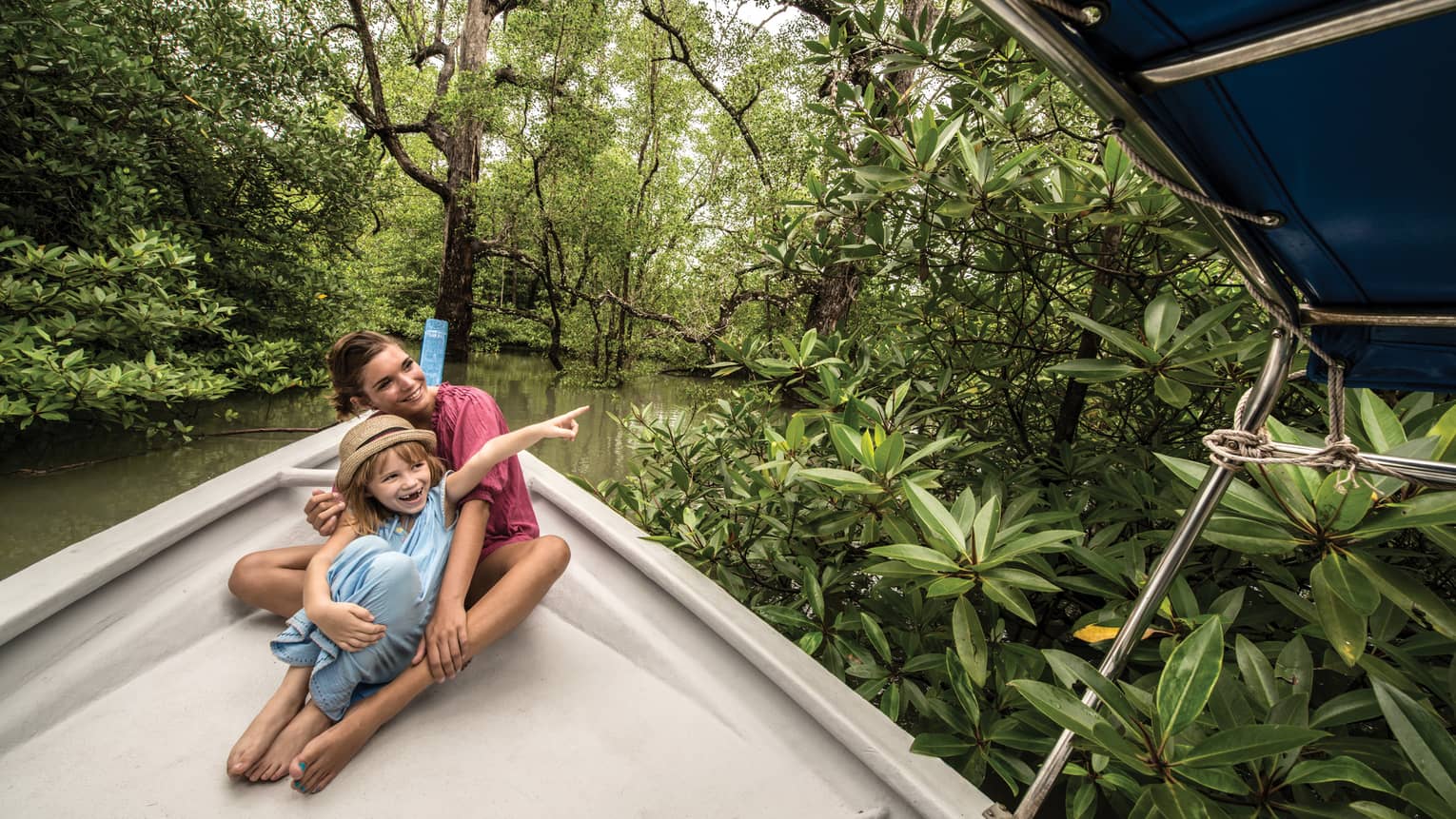 A woman and child on the bow of a canopied riverboat amid lush, green mangrove trees; both gaze where the child is pointing.