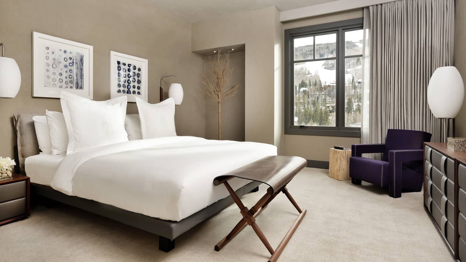 Bed with white linens, folding bench at foot, armchair by window with mountain views