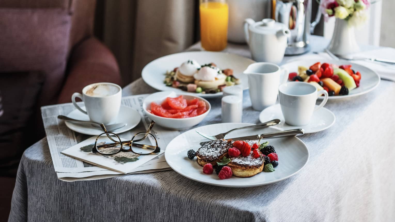 Breakfast dishes on white round dishes, square white table, orange juice, coffee, eyeglasses