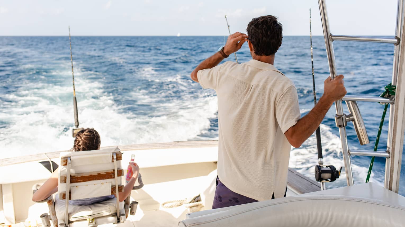Two guests on a boat gaze at the foamy wake behind the craft, which has multiple upright fishing rods attached to its sides.